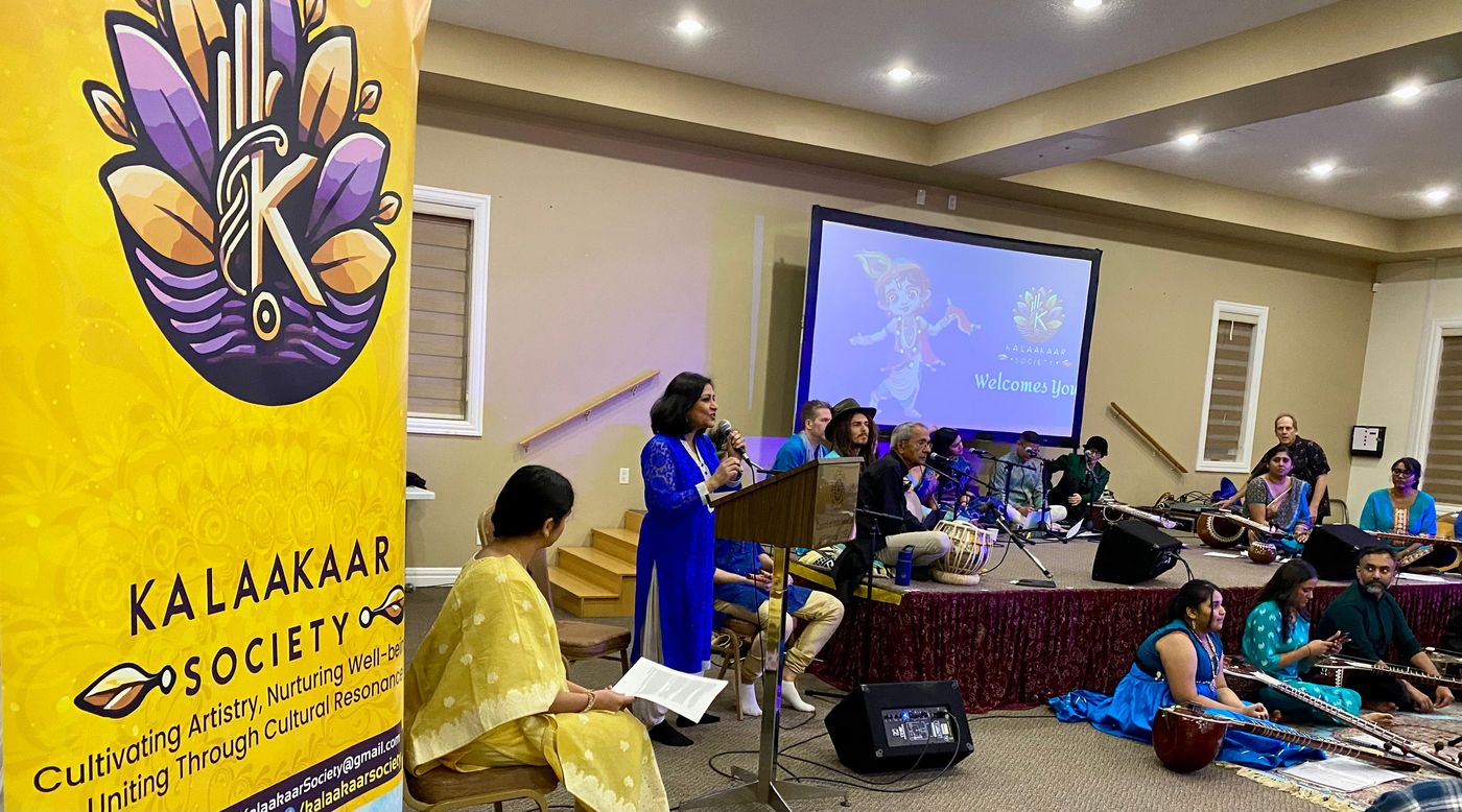 A speaker welcoming the audience at Chai and Chant — Krishna, with musicians on stage behind her